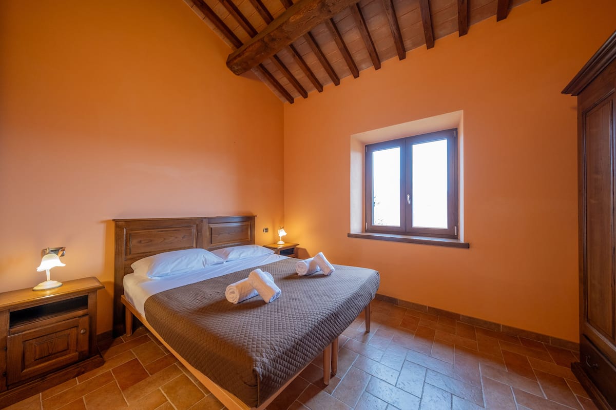Un dormitorio con una cama de madera, una mesita de noche con lámpara, una ventana y un armario.