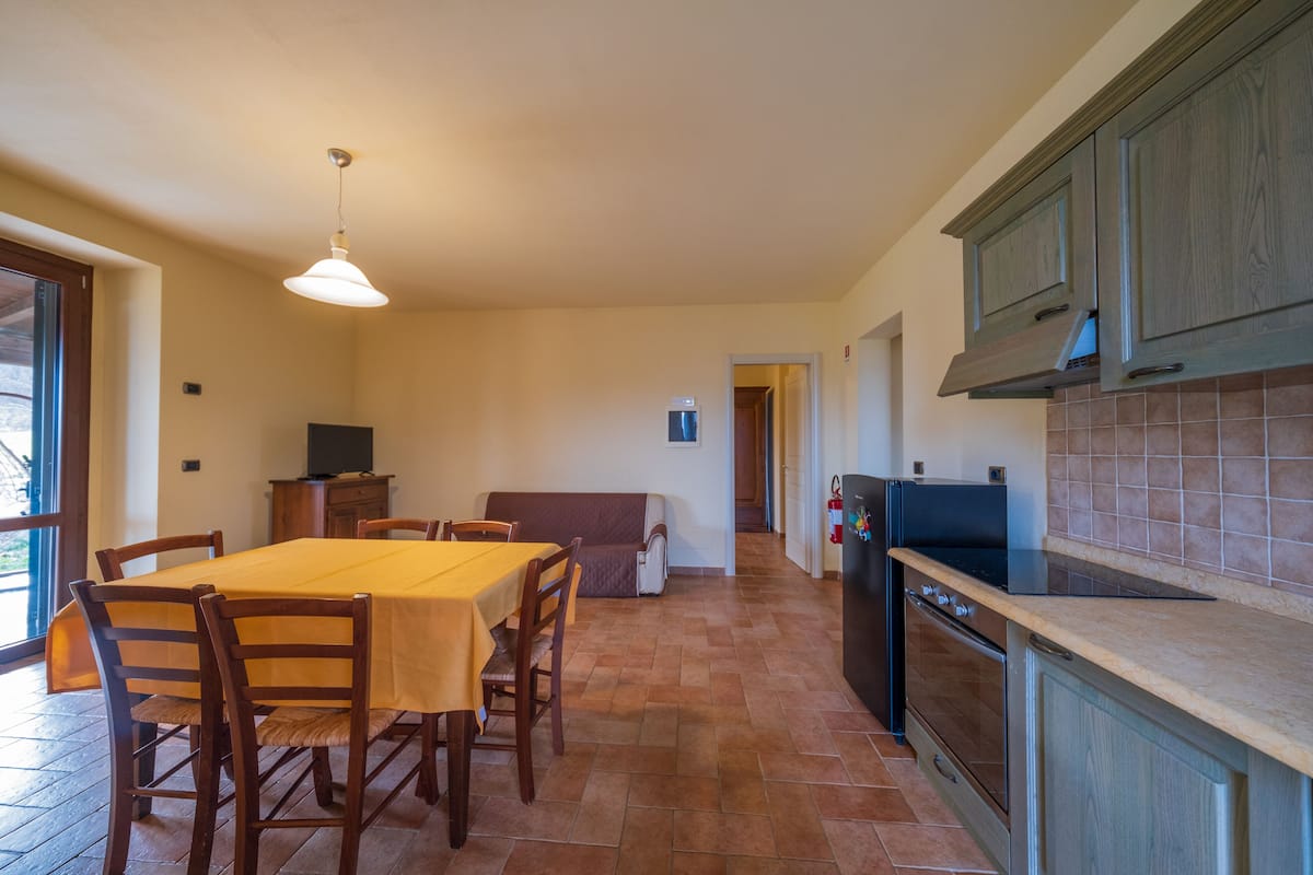 Un comedor con mesa y sillas, una cocina con armarios y electrodomésticos, y vista al exterior por una puerta.