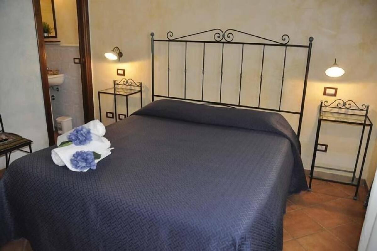 Un lit bien fait, recouvert d’une couvre-lit bleu foncé, et agrémenté d’une seule fleur décorative. Il y a deux tables de chevet avec des lampes, ainsi qu’une petite table sur laquelle est posée une plante. La pièce présente un aménagement simple et épuré, avec un sol carrelé.