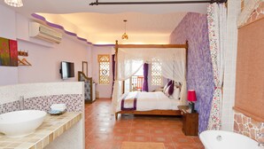 Un dormitorio con cama con dosel, un ventanal con cortinas moradas, una mesita de noche con lámpara y vistas al balcón exterior.