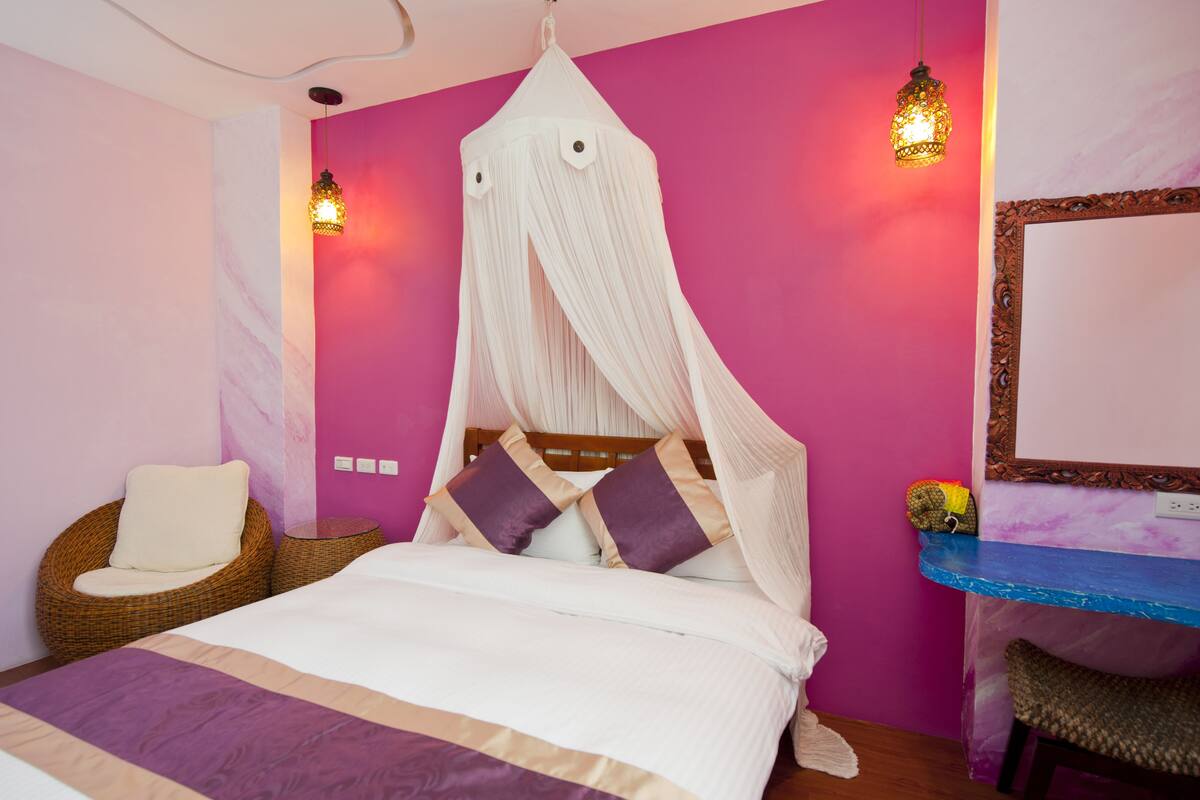 Una habitación con una cama, una silla de mimbre, una mesita y un espejo en la pared.