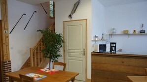 Une cuisine compacte équipée d’un four à micro-ondes, d’une machine à café et d’étagères. Une table à manger en bois, avec des chaises, se trouve au premier plan, et un escalier agrémenté d’une plante est visible.
