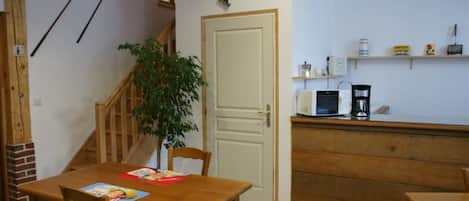 Une cuisine compacte équipée d’un four à micro-ondes, d’une machine à café et d’étagères. Une table à manger en bois, avec des chaises, se trouve au premier plan, et un escalier agrémenté d’une plante est visible.