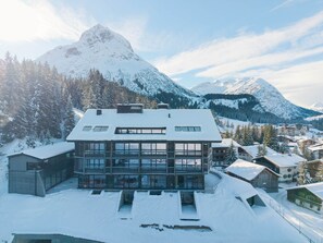 A modern multi-story building with large windows, surrounded by snow-covered mountains and trees. - New Hotel Kristberg in Lech with Spa, outdoor pool and panoramic view (Lech am Arlberg)