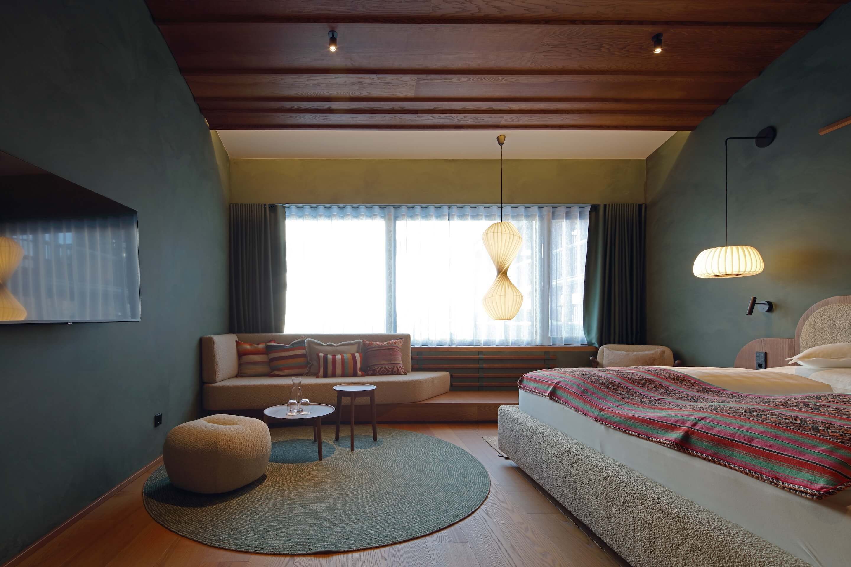 Quarto de hotel moderno com teto de madeira, uma cama grande com manta estampada, sofá com almofadas, um banquinho redondo pequeno e um tapete redondo.