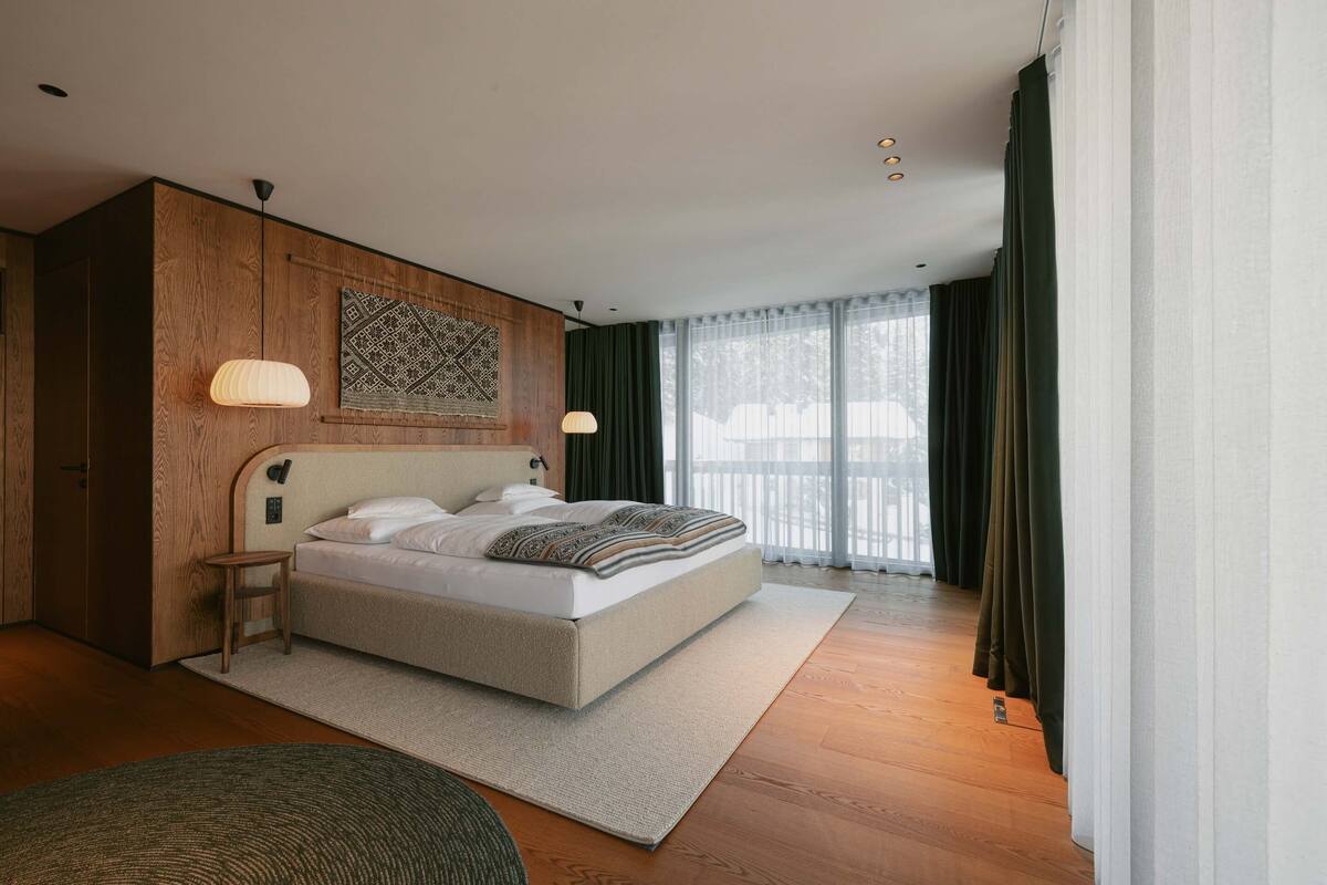 Une chambre moderne avec un grand lit, un lambris en bois, une petite table de chevet et une vue sur l’extérieur grâce à de grandes fenêtres.
