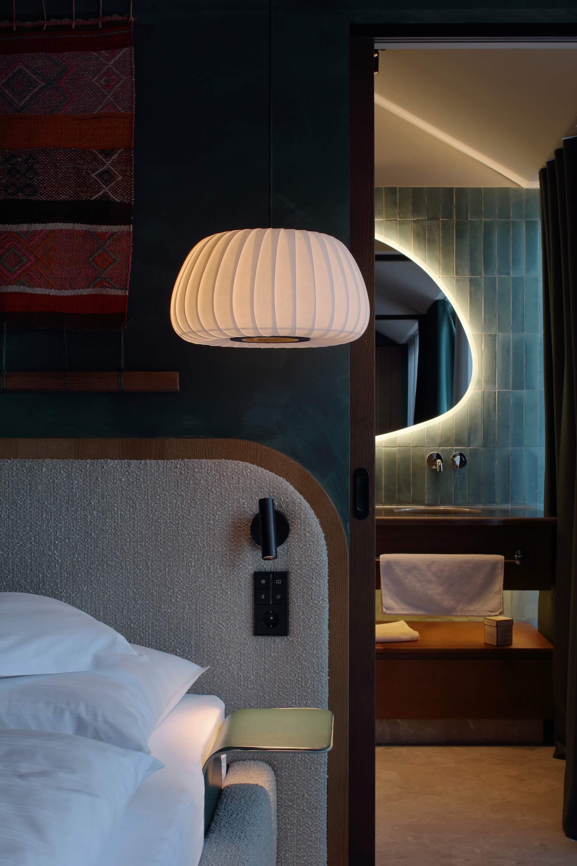 Habitación de hotel con cama, mesita de noche, lámpara, baño con lavamanos y espejo, y un tapiz decorativo en la pared.