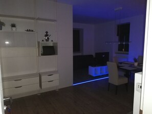 Een moderne woonkamer met een eetgedeelte, met een wit kastje, een televisie en een blauwe LED-lichtstrip.