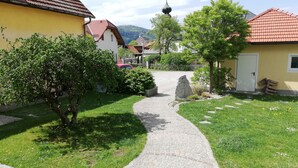 A cobblestone pathway leading to a yellow building with a red-tiled roof, surrounded by greenery and a distant church spire.