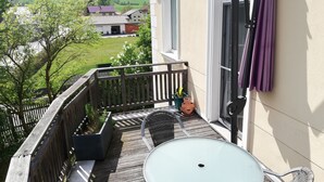 A balcony with a table and chairs, a potted plant, and a view of a green landscape with houses.