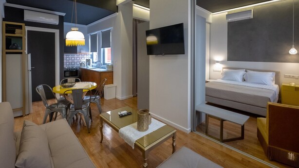 A modern living room with a sofa, a coffee table, and a dining area with chairs. There is a TV mounted on the wall and a bed with a bench in the adjacent room. - Athenian Yard Suites (Athens)
