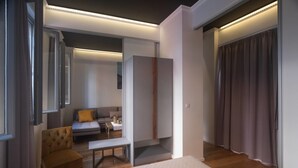 A modern hotel room with a bed, a seating area with a sofa and armchair, a small table, and a wardrobe. - Athenian Yard Suites (Athens)