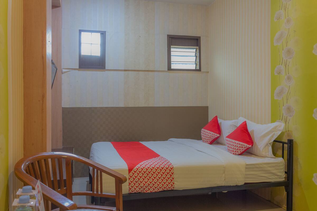 A single bed with a red and white patterned bedspread, two pillows, a wooden chair, and a window above the bed.