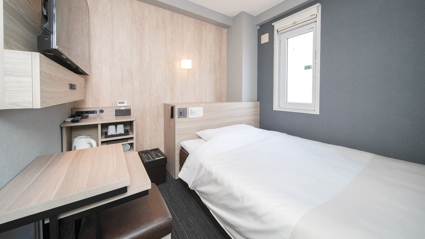 A compact hotel room with a bed, a desk, a chair, a small table, a trash bin, and a window.