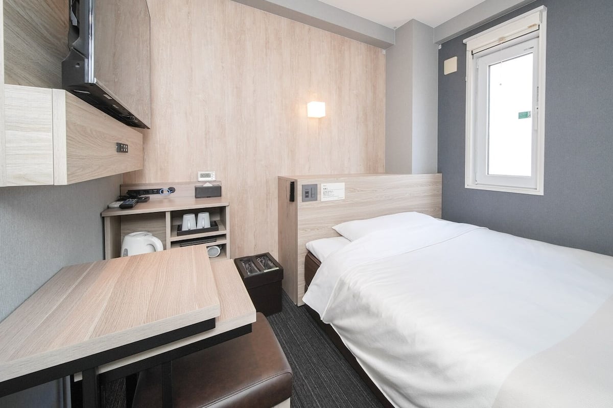 A compact hotel room with a bed, a desk, a chair, a small table, a trash bin, and a window.