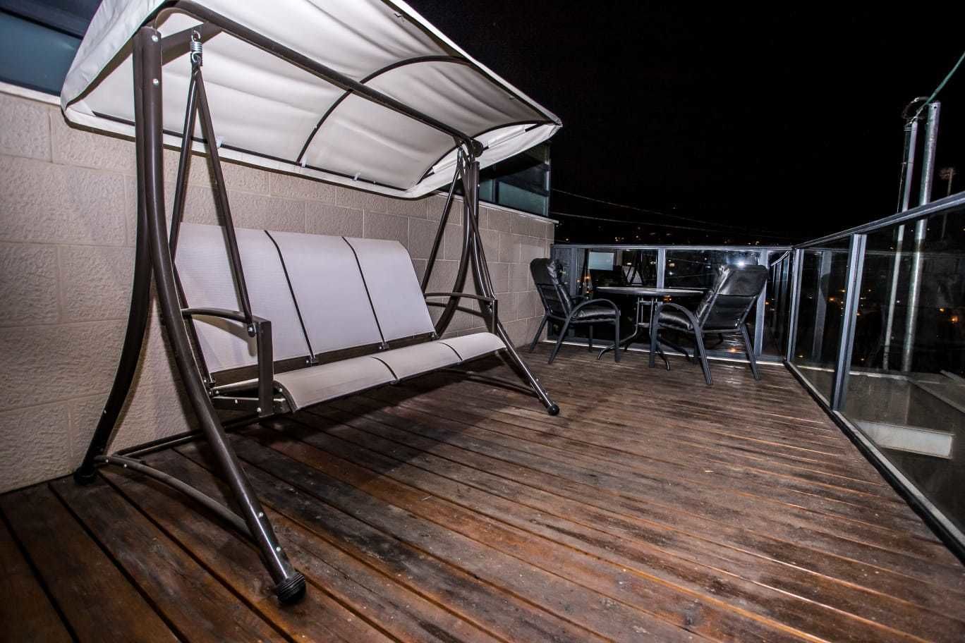 A wooden deck with a swing chair and a table with chairs, overlooking a cityscape at night.