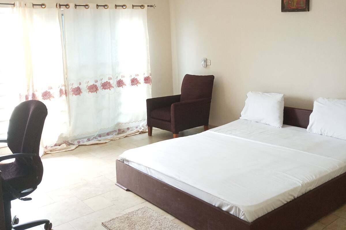 A bedroom with a large bed, a desk, a chair, a ceiling fan, and a window with curtains.