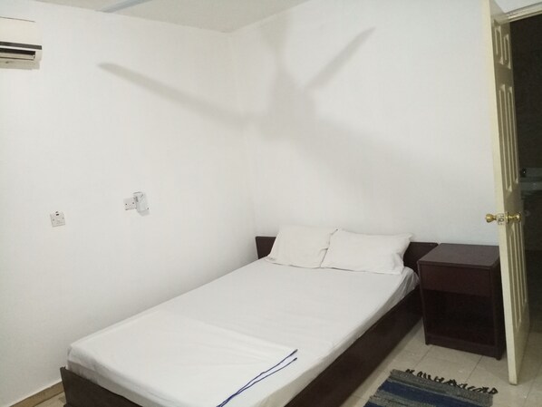 A small, single-bed room with a white bedspread, a wooden nightstand, and a wall-mounted air conditioning unit.