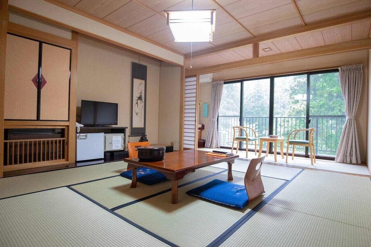 Una tradizionale stanza giapponese con pavimento in tatami, un tavolino, una televisione e un balcone con sedie.