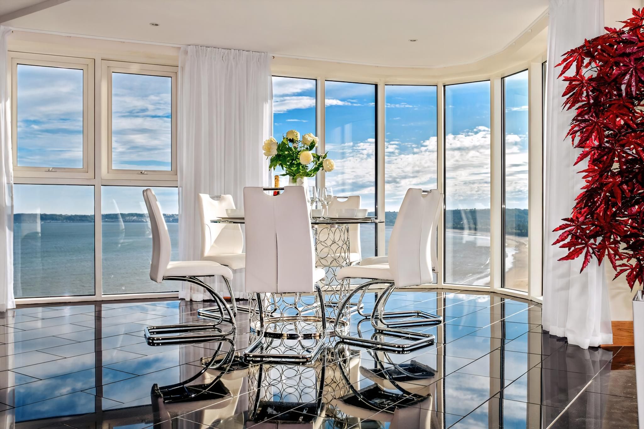 A modern dining area with a glass table and chairs, a vase of flowers, and a view of the water.