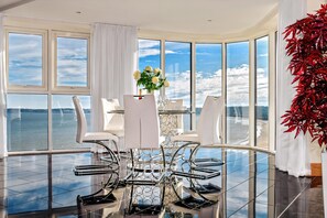 A modern dining area with a glass table and chairs, a vase of flowers, and a view of the water. - Just Stay Wales – Meridian Quay Penthouses (Swansea)