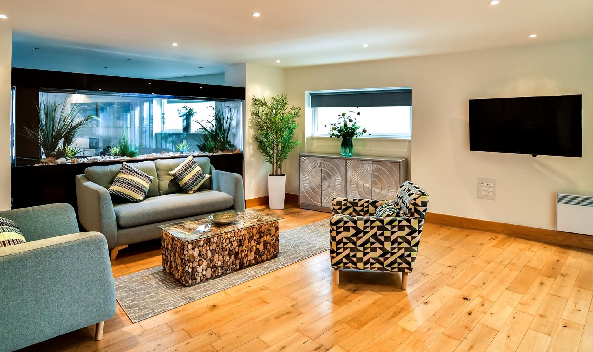 A modern living room with a sofa, patterned armchair, wooden coffee table, and a large fish tank.