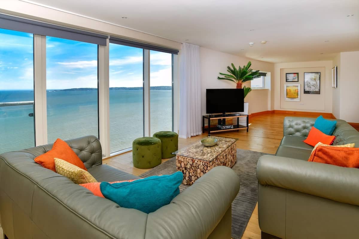 A modern living room with a large window overlooking a body of water, a flat-screen TV, and a green plant.