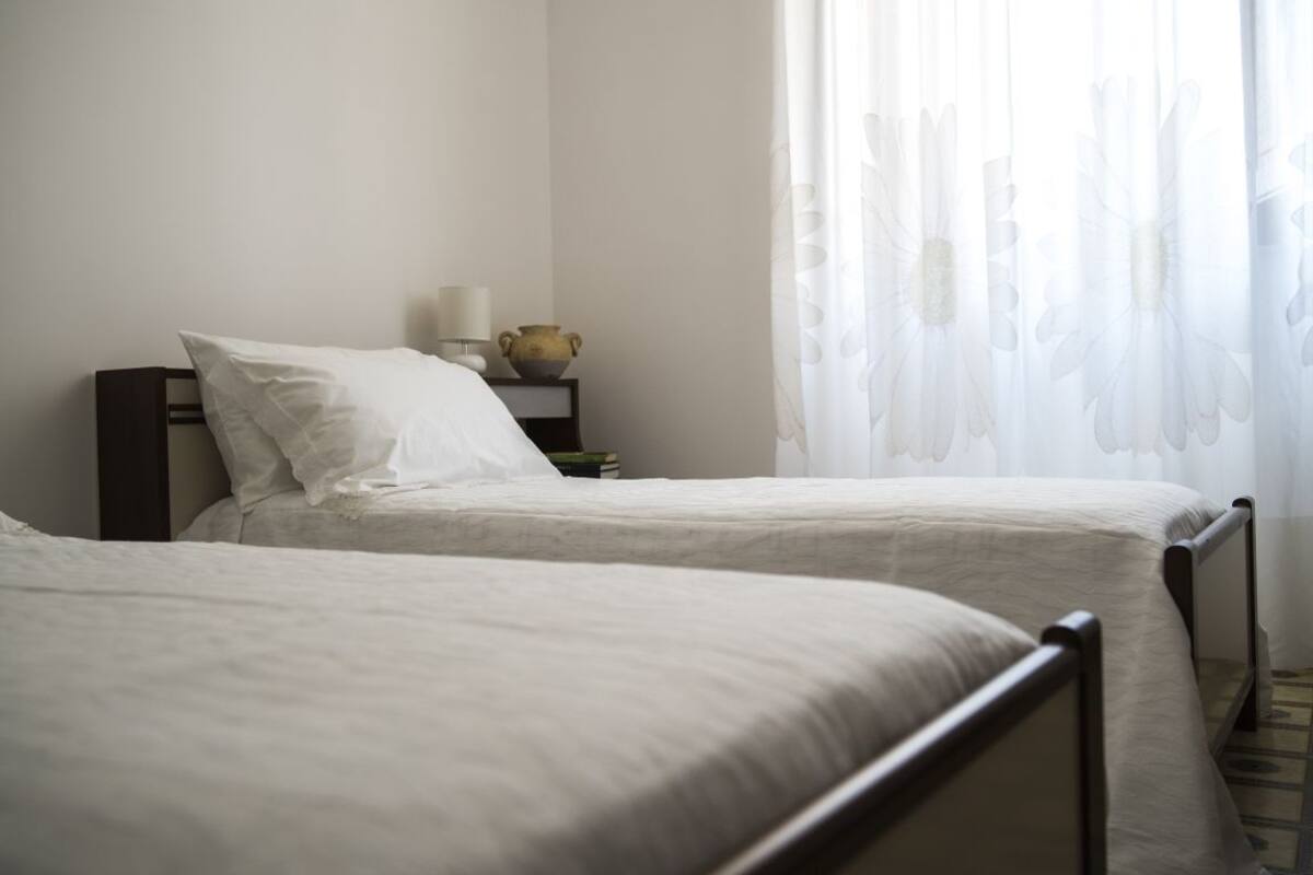 Une chambre avec deux lits simples, une table de chevet avec une lampe et un vase, et des rideaux légers.