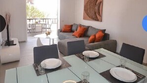Un espace repas moderne avec une table en verre pour quatre personnes, un canapé avec des coussins orange et une grande œuvre d’art abstraite accrochée au mur.