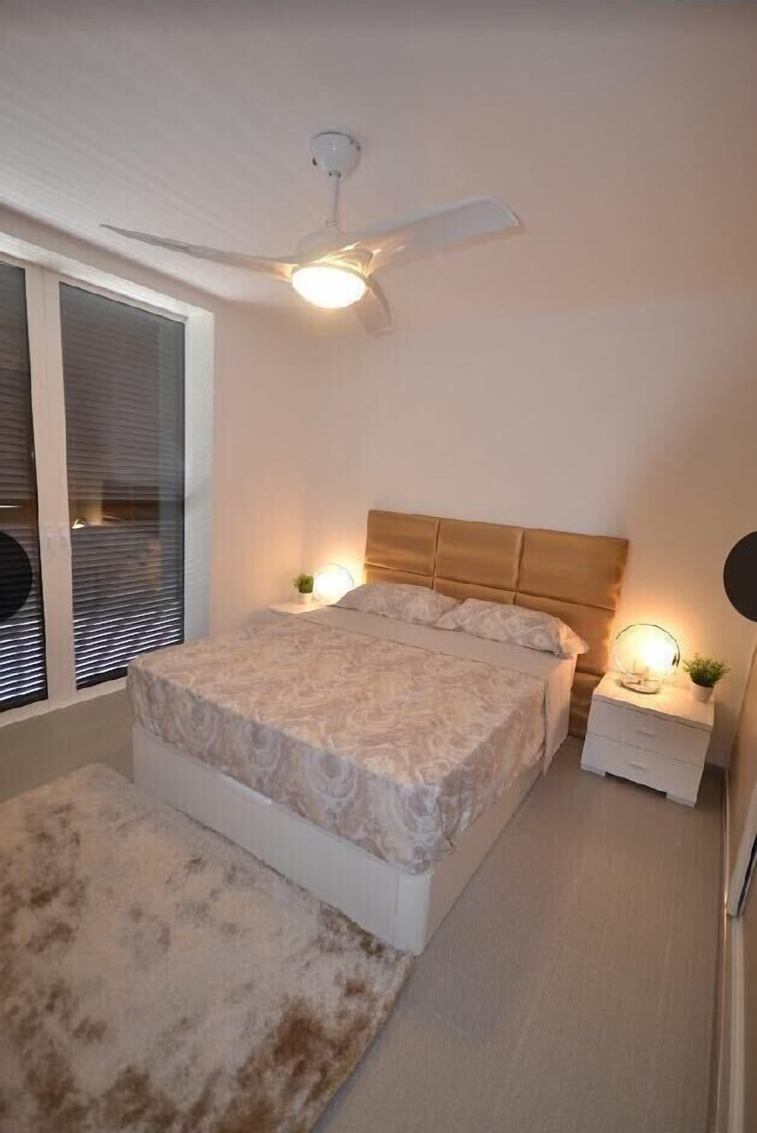 Un dormitorio con cama, mesitas de noche, ventilador de techo y ventana con persianas.