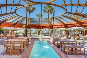 An outdoor pool area with wooden tables and chairs, surrounded by palm trees and a covered seating area.