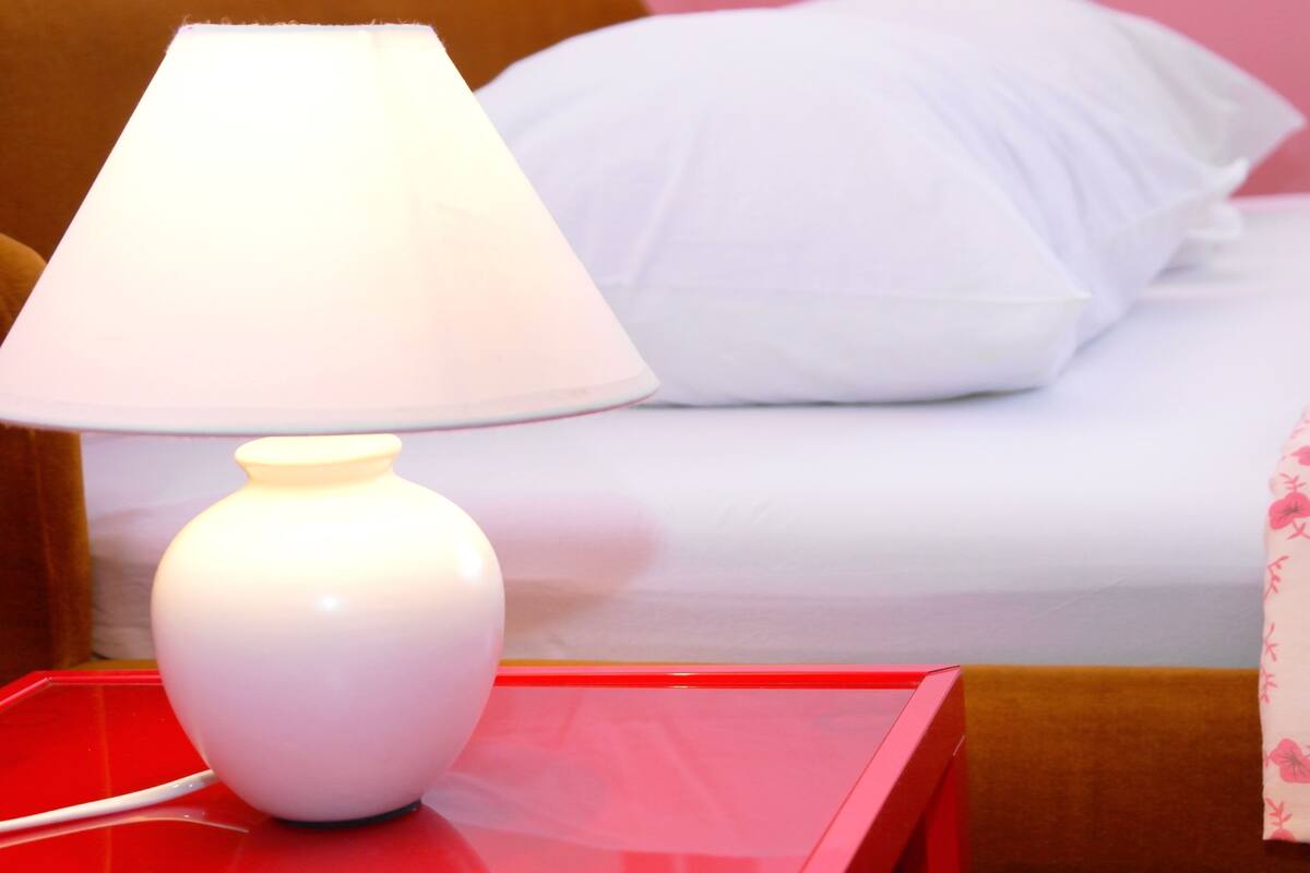 A bed with white bedding, a white lamp on a red nightstand, and a floral-patterned pillow.