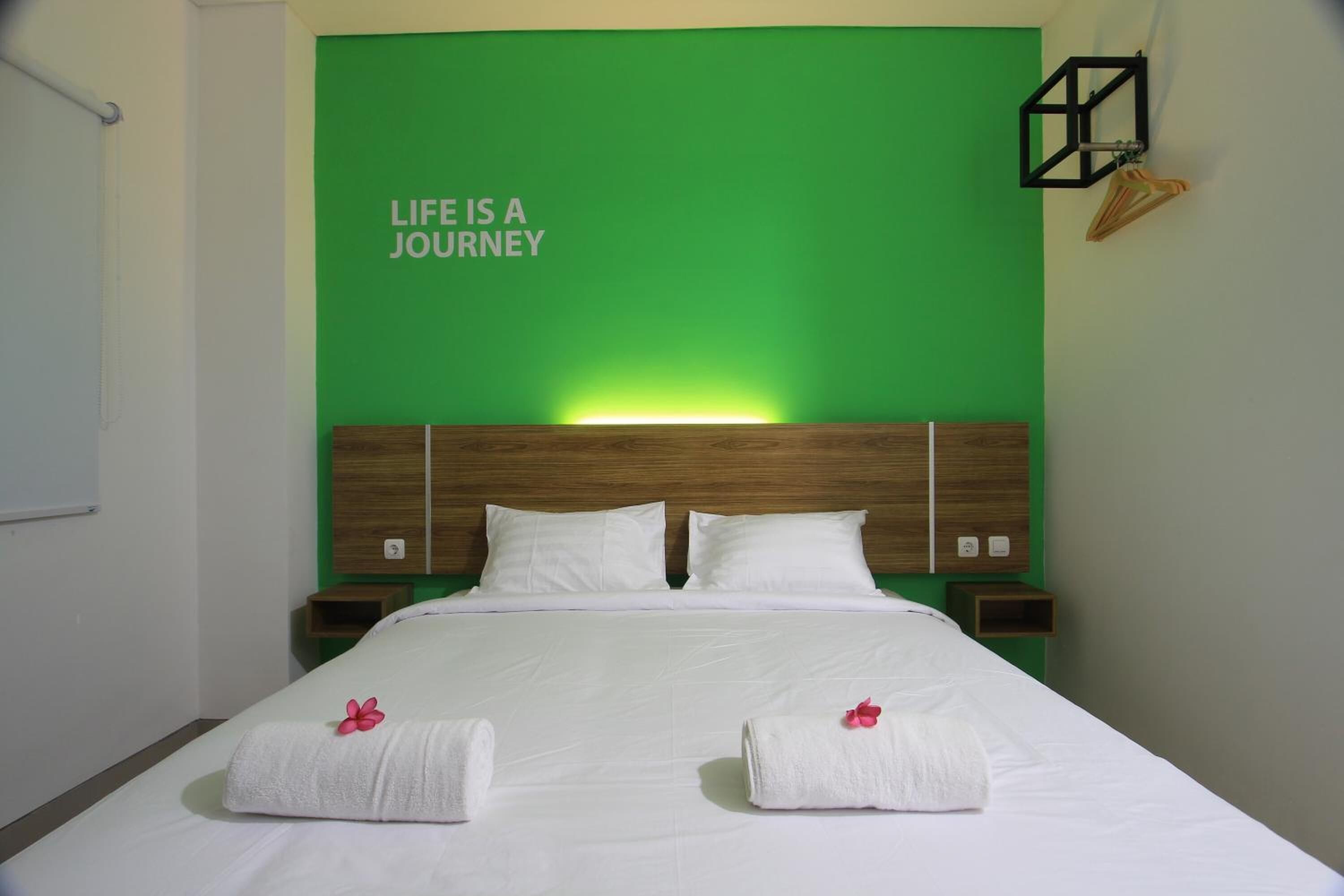A hotel room with a bed, two white towels with pink flowers, a green wall with the text "LIFE IS A JOURNEY", and a wooden headboard.