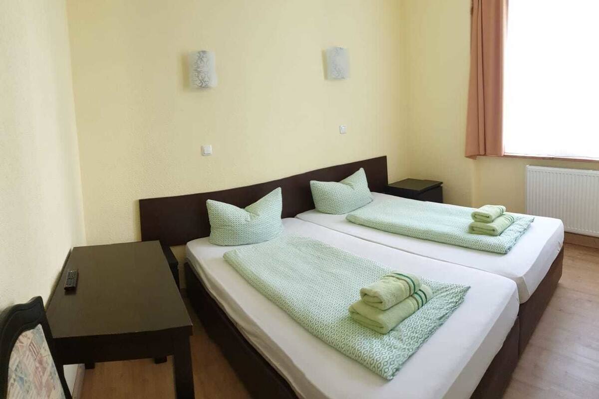 Habitación de hotel con dos camas, cada una con una manta y almohada verdes, una mesita y una silla.
