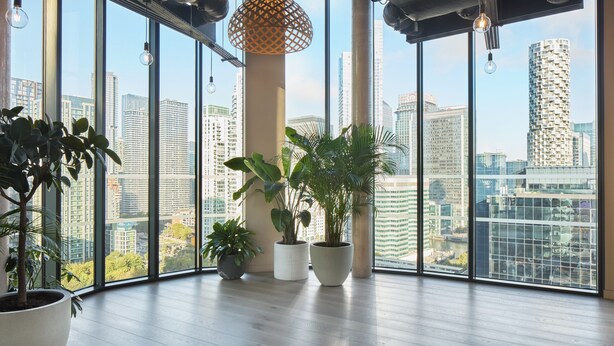 A spacious room with large windows offering a city view, wooden flooring, and several potted plants. - ARK Canary Wharf (London)