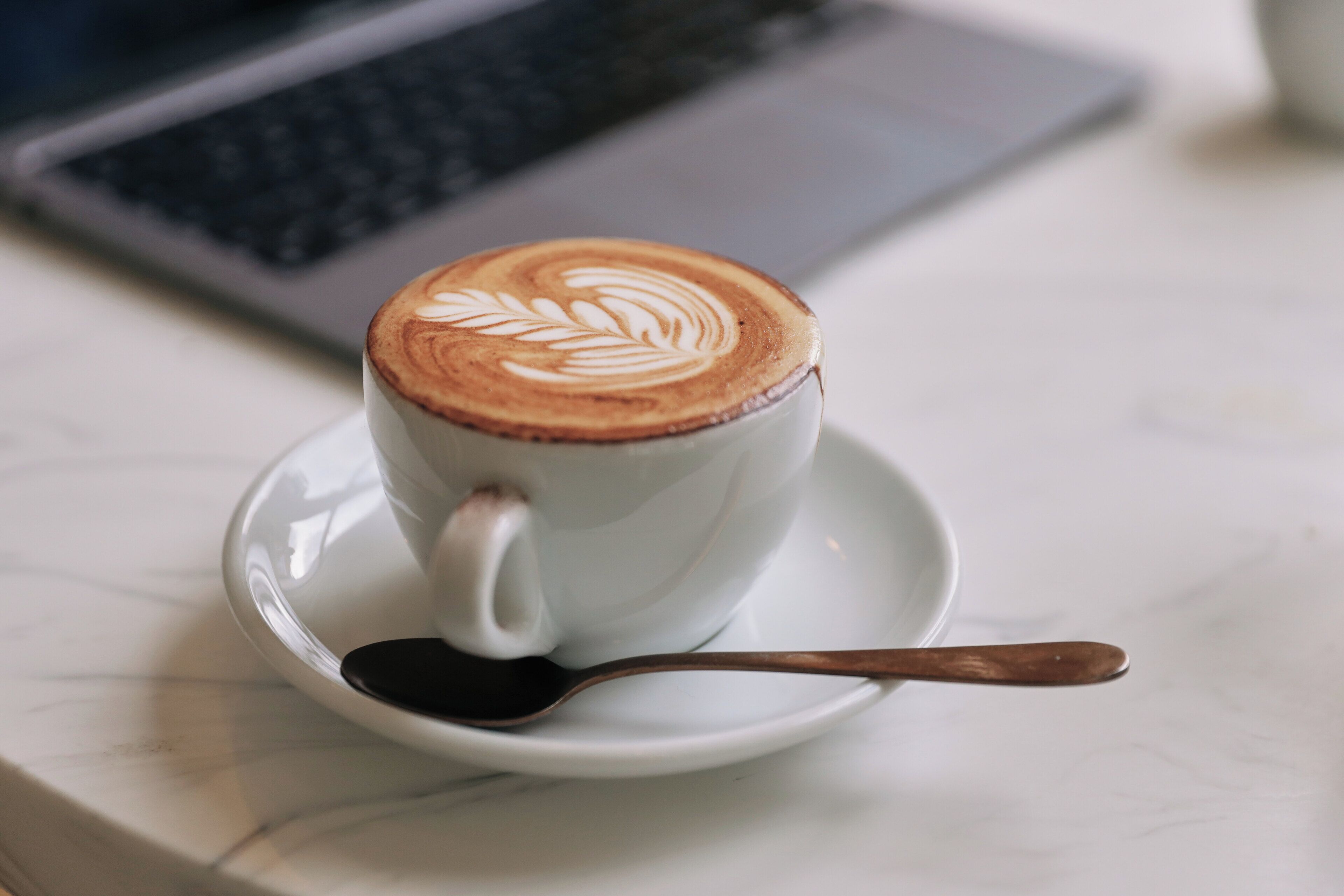 Eine Kaffeetasse mit Latte-Art auf einem Untertasse, platziert auf einer Marmorplatte, im Hintergrund ein Laptop.