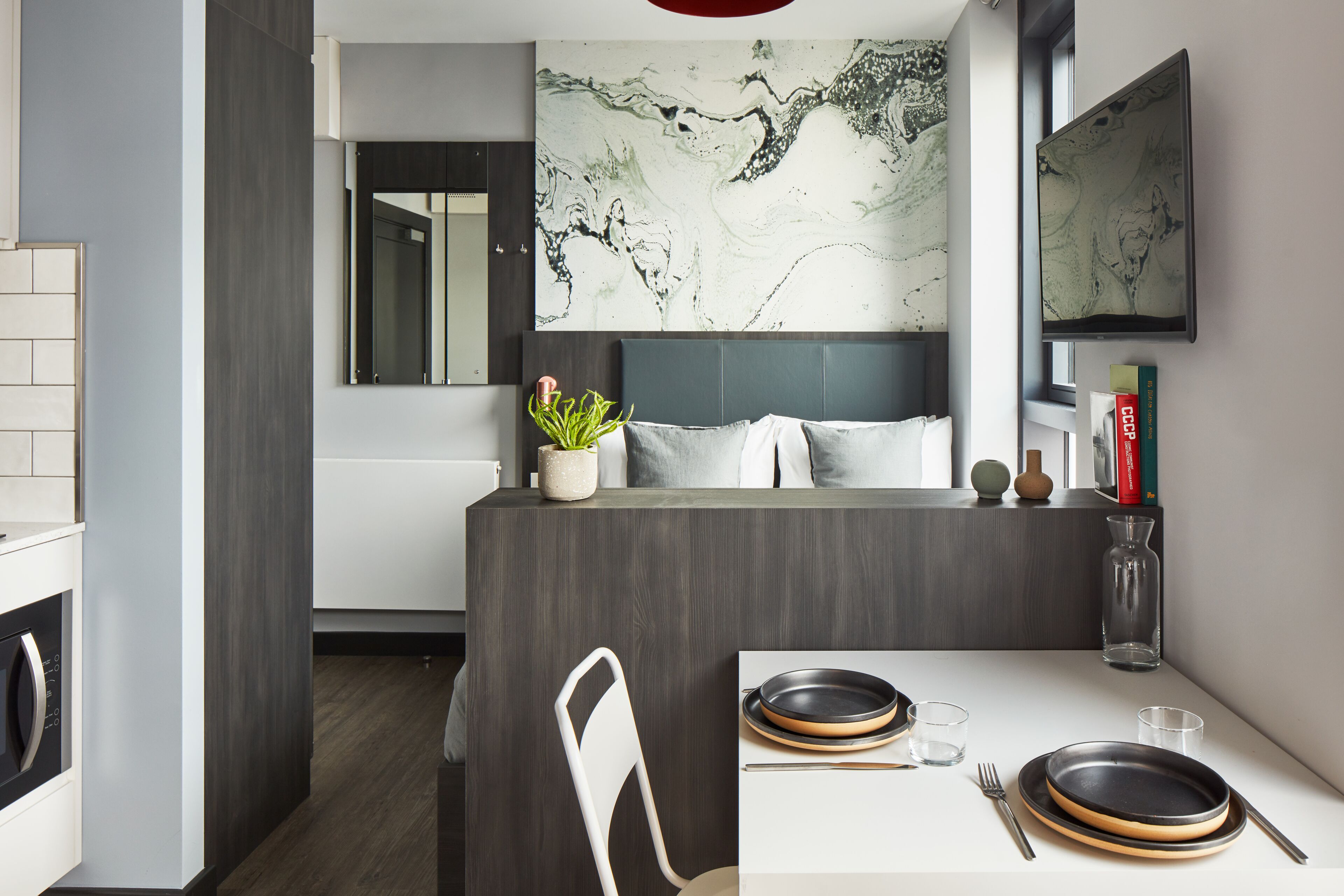 Una cocina moderna con salpicadero de mármol, un comedor con mesa blanca para dos comensales y un cabecero gris al fondo.