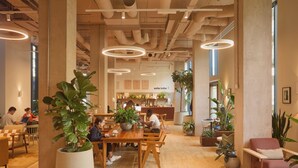 A spacious cafe with wooden tables and chairs, large potted plants, and a modern industrial ceiling design. - ARK Canary Wharf (London)