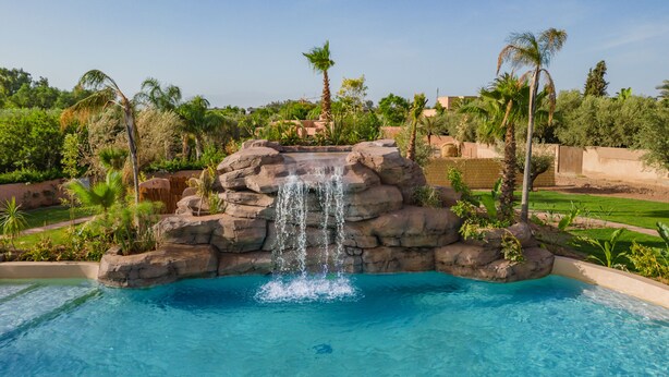 A pool with a waterfall feature surrounded by palm trees and greenery. - Oasis Lodges (Marrakech)