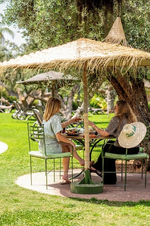Una persona cenando bajo un toldo de paja en una mesa al aire libre, con sillas y una mesa de comedor a la vista.