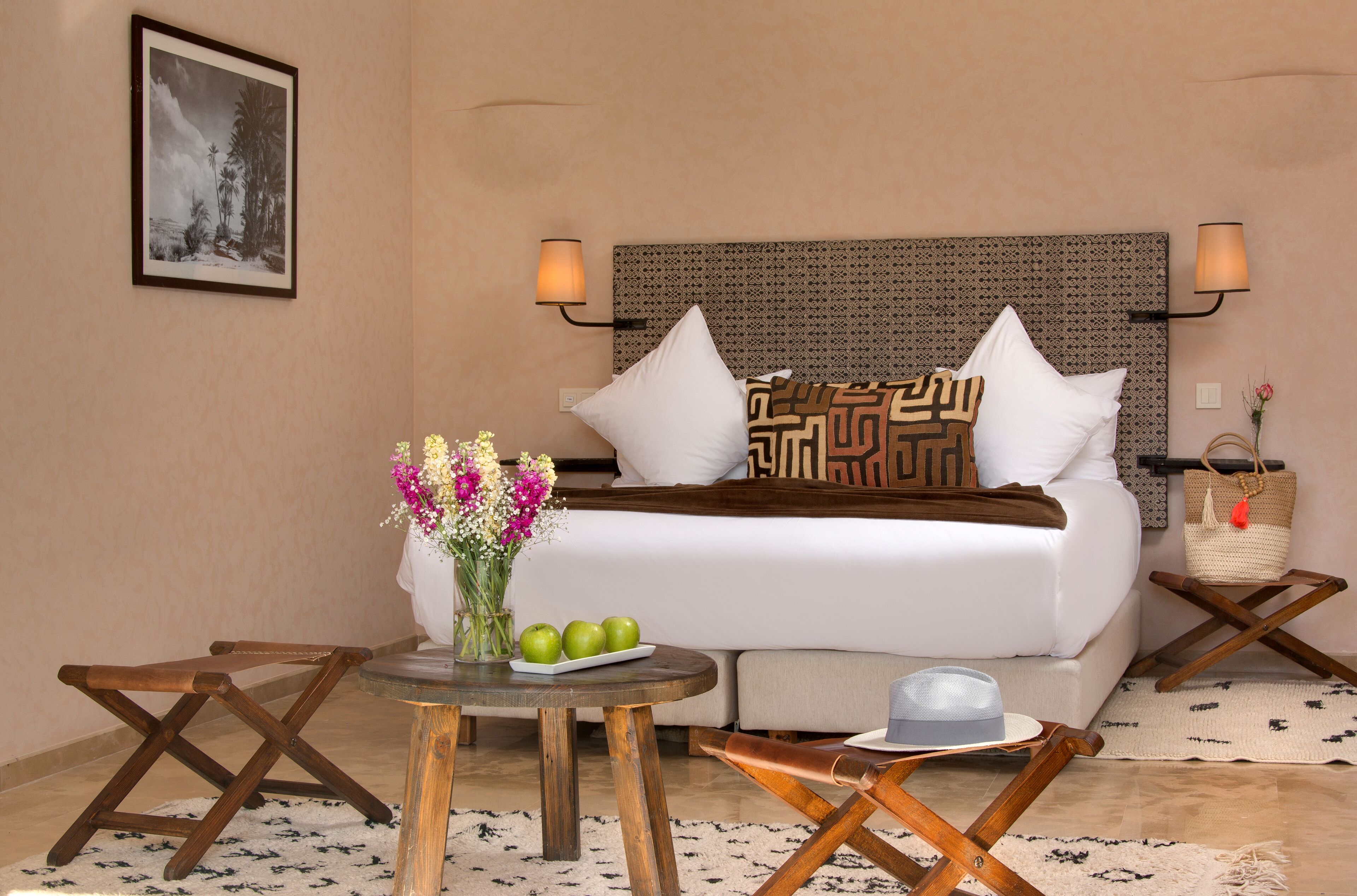 Habitación de hotel ordenada con una cama, dos lámparas de noche, una mesita con un jarrón de flores y manzanas, y una cesta tejida en una mesita auxiliar.