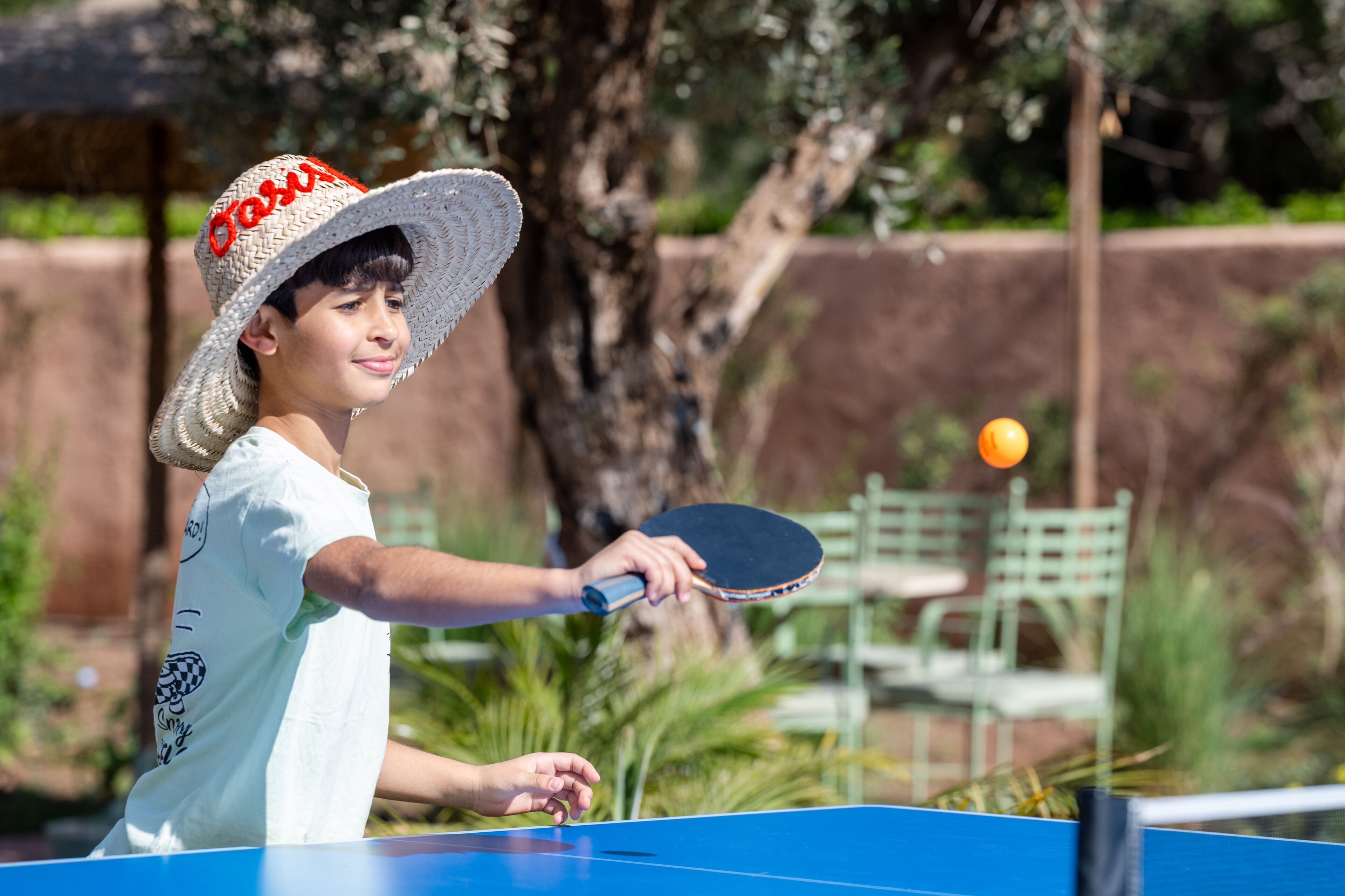 A person playing table tennis outdoors with a blue table and a green chair in the background.