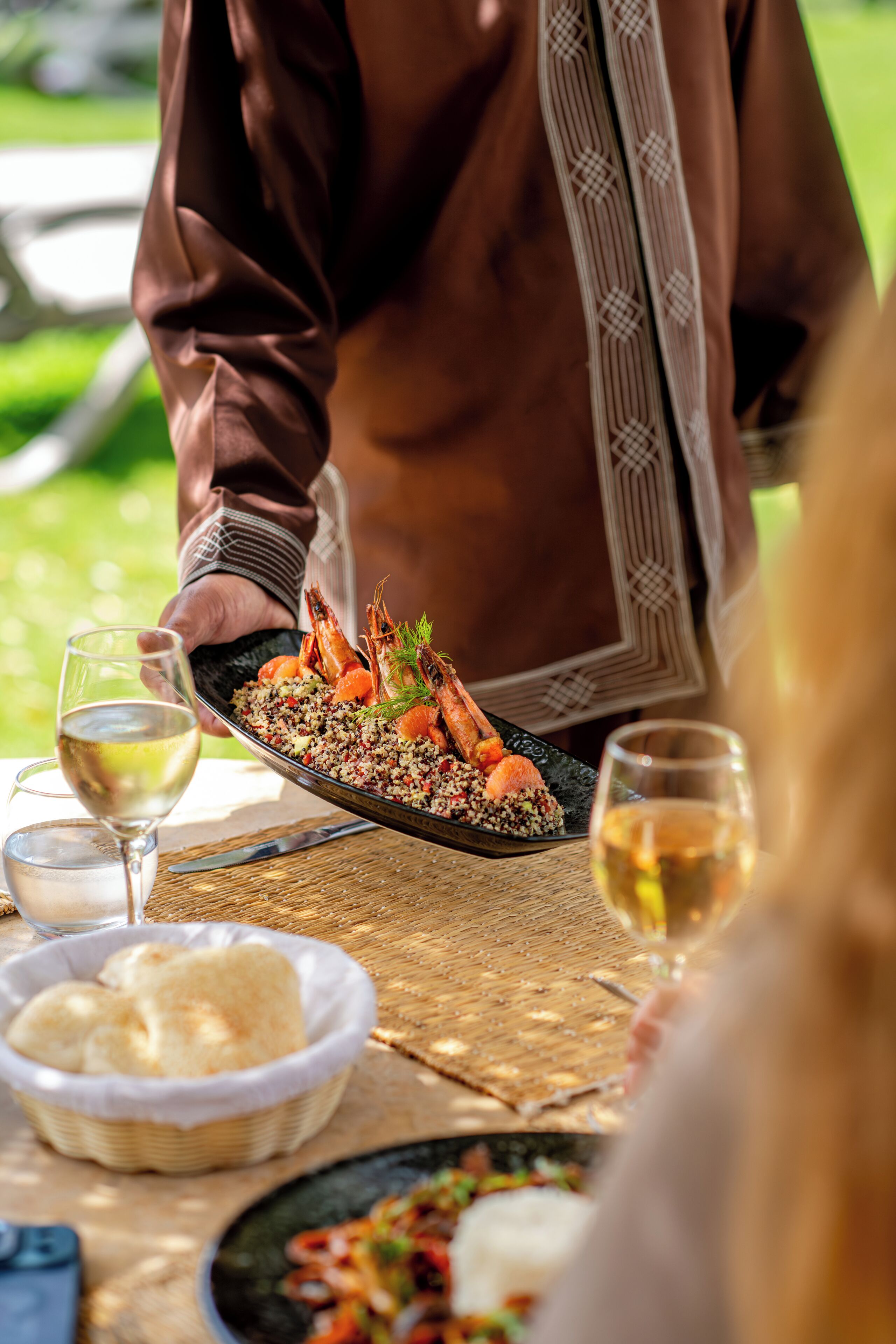 A dining table with a plate of food, a glass of white wine, and a basket of bread.
