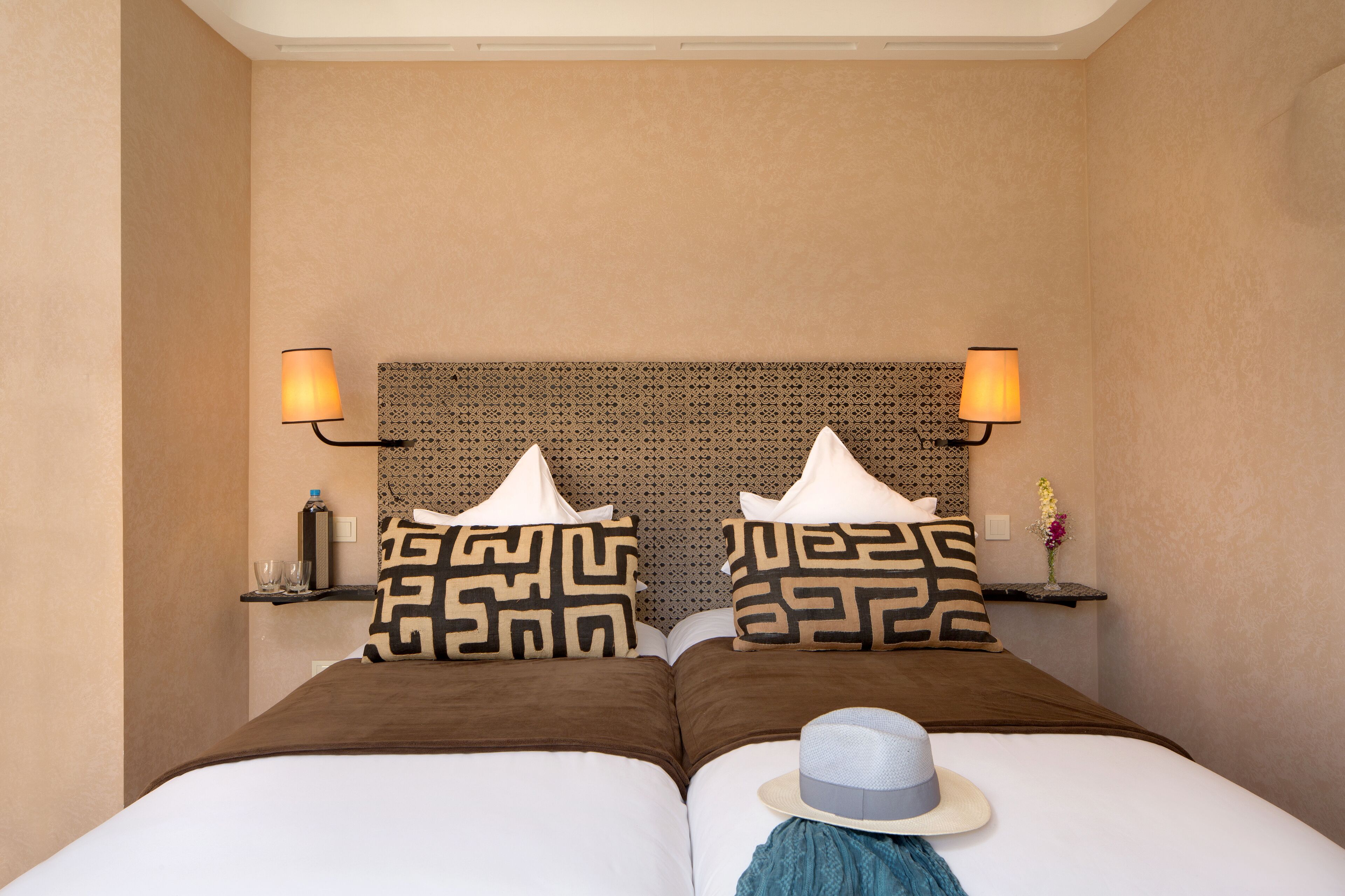 A hotel room with two beds, patterned pillows, bedside lamps, a small shelf with a vase, and a hat on one bed.