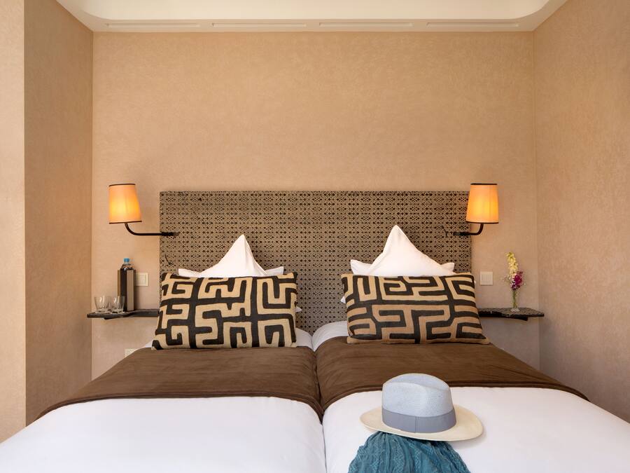 A hotel room with two beds, patterned pillows, bedside lamps, a small shelf with a vase, and a hat on one bed.