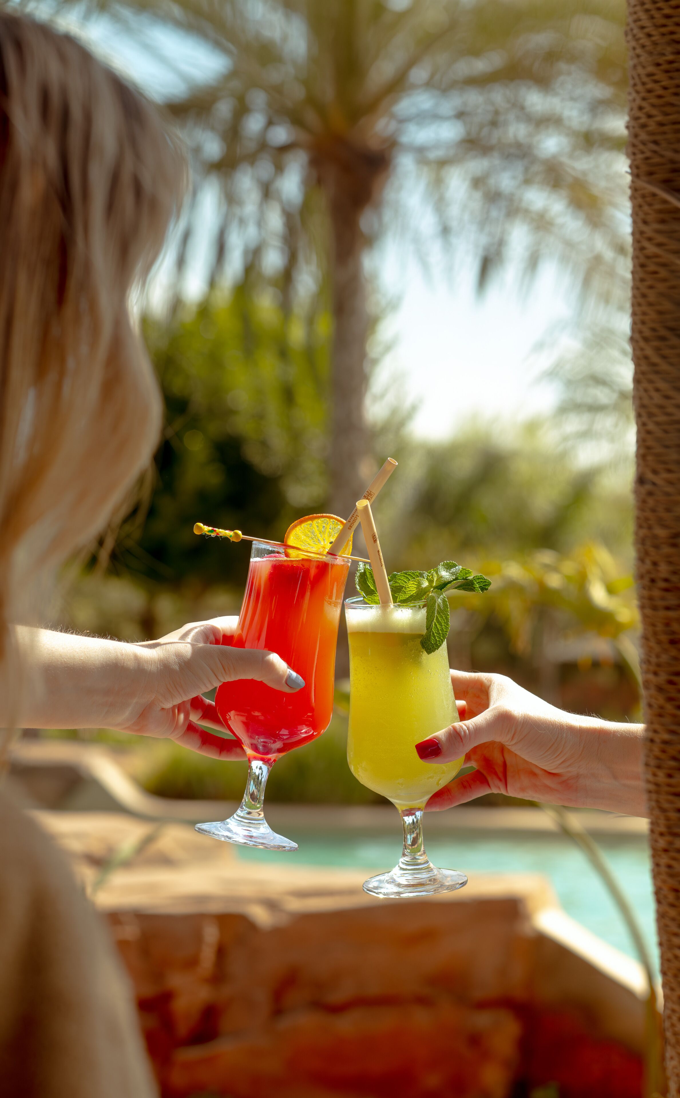 Twee personen houden cocktailglazen met garnering vast, buiten met een zwembad en palmbomen op de achtergrond.