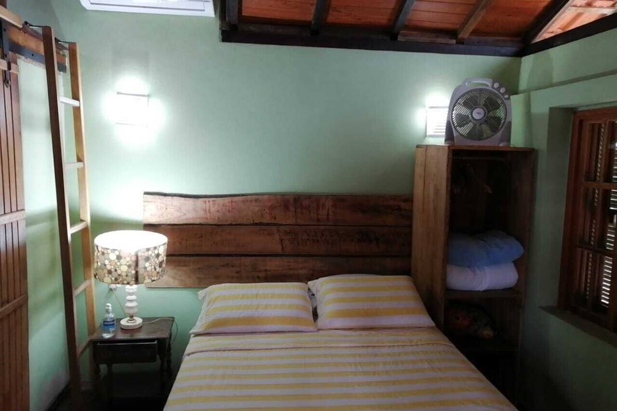 A bedroom with a wooden headboard, a bed with striped bedding, a bedside lamp, a fan, and a shelf with pillows.