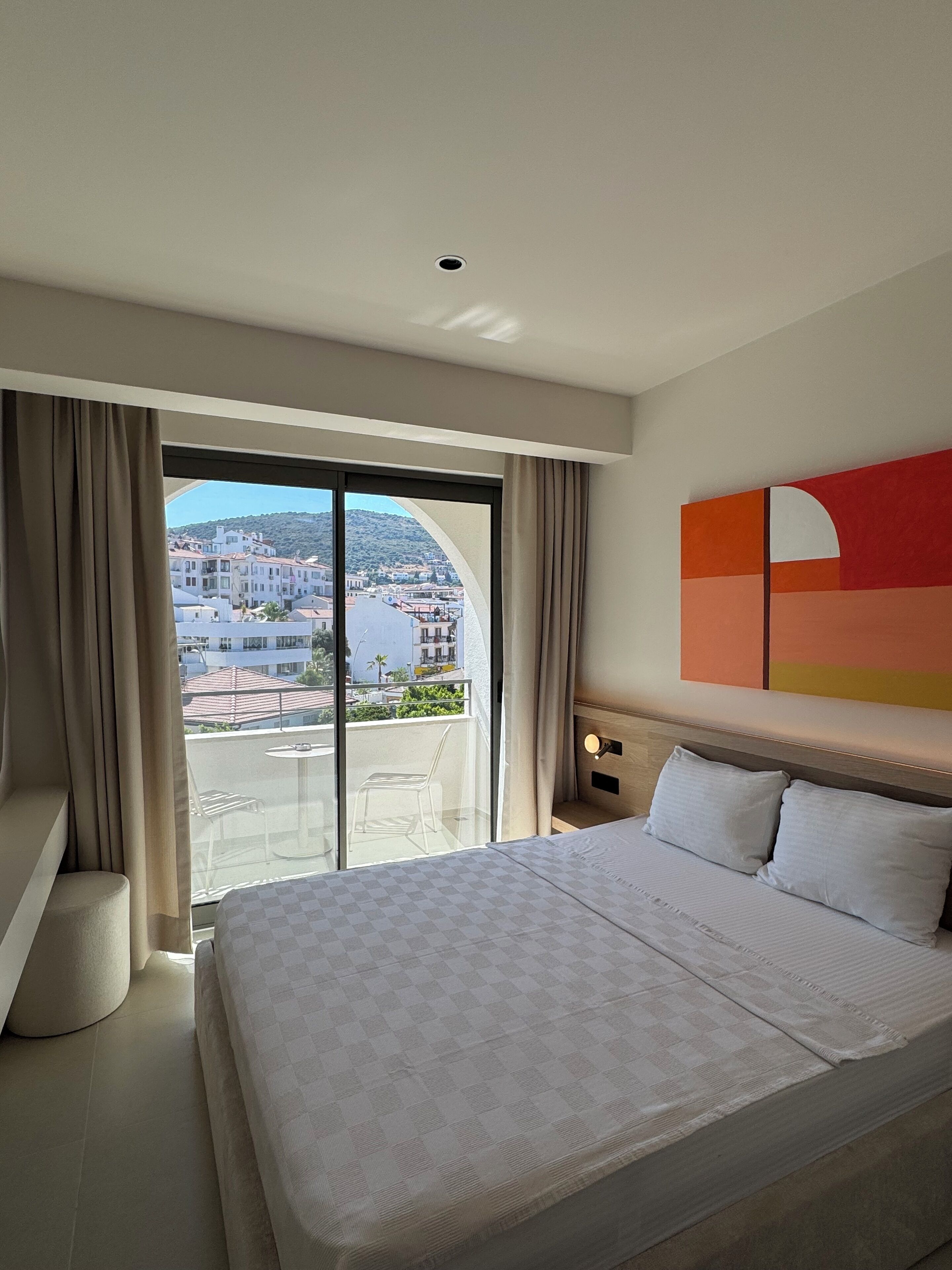 A hotel room with a large bed, a view of buildings through a sliding glass door, and a modern wall art piece.