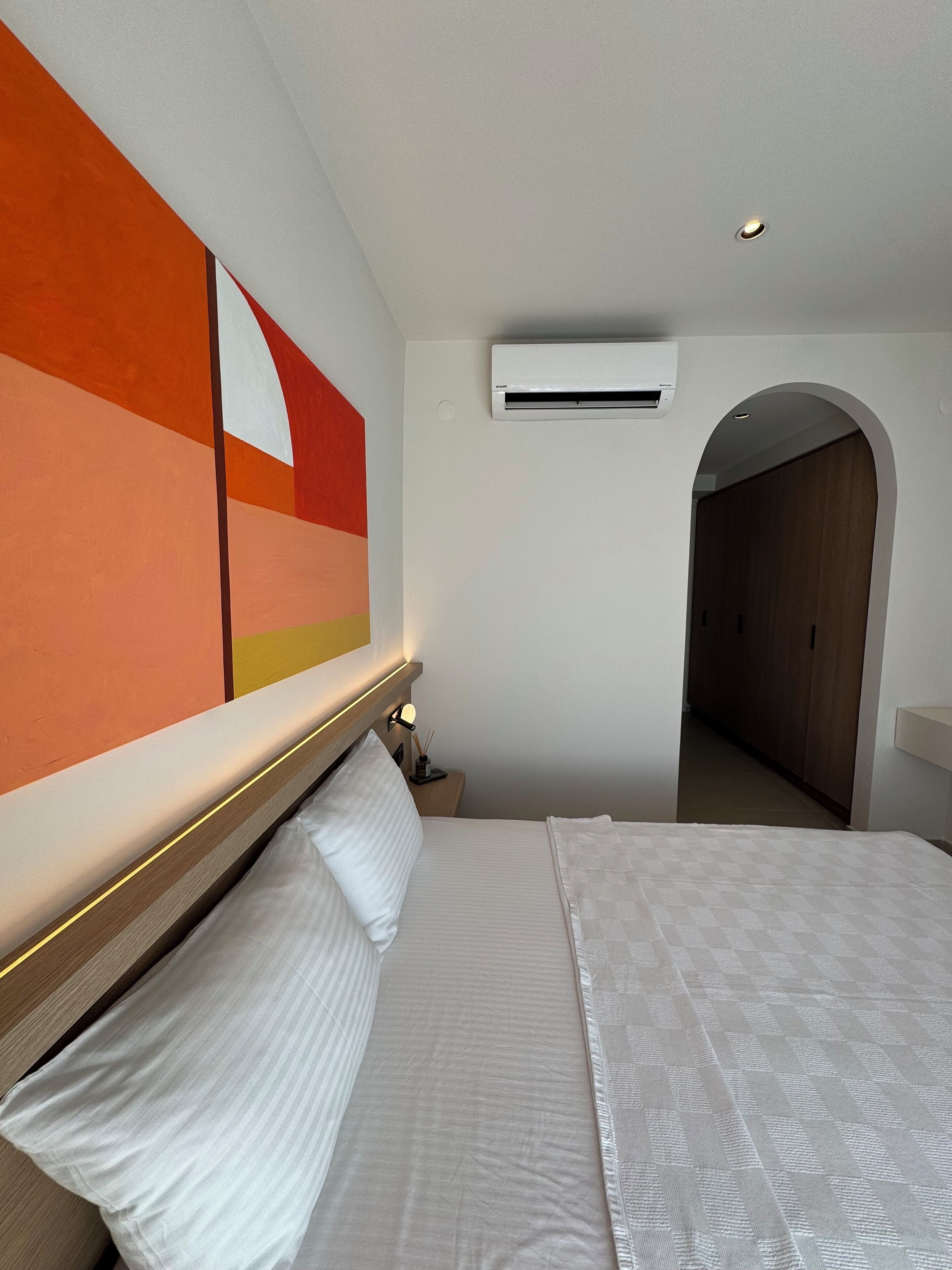 Una habitación de hotel con una cama grande, un equipo de aire acondicionado, una mesita de noche con lámpara y un pasillo que conduce a una puerta de madera.