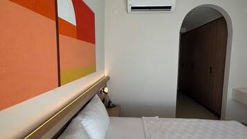 Una habitación de hotel con una cama grande, un equipo de aire acondicionado, una mesita de noche con lámpara y un pasillo que conduce a una puerta de madera.